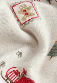 Cremefarbenes Gewebe mit gestickten Feiertagsmotiven, darunter ein Lebkuchenmann, Bäume und ein Schlitten mit Geschenken in Rot und Grün.