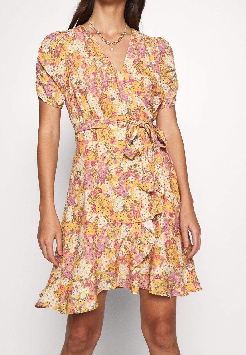 Robe portefeuille florale jaune avec un ourlet à volant et des manches bouffantes, nouée à la taille, portée avec un collier en or à plusieurs couches.