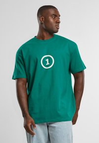 Camiseta de algodón verde con un corte relajado, que presenta un gráfico circular blanco con el número "1" en el centro. Manga corta y escote redondo.