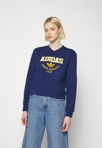 Tamsiai mėlynas megztinis su v-kaklu, su geltonu "ADIDAS ATHLETIC CLUB" logotipu ir ikoniniu lapų dizainu. Minkšta medžiaga, laisvas kirpimas.