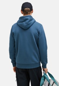 Plava dukserica od mekanog materijala, s kengur džepom i podesivom kapuljačom. Model drži sivu sportsku torbu s tirkiznim trakama.