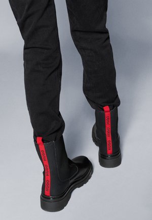 Botas pretas até ao tornozelo com tecido texturizado e sola de borracha. Acabamentos em vermelho com o logótipo da marca na parte de trás; acabamento brilhante na ponta.