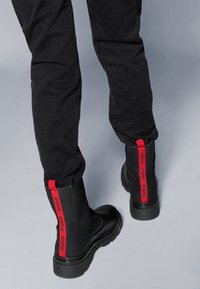 Botas pretas até ao tornozelo com tecido texturizado e sola de borracha. Acabamentos em vermelho com o logótipo da marca na parte de trás; acabamento brilhante na ponta.