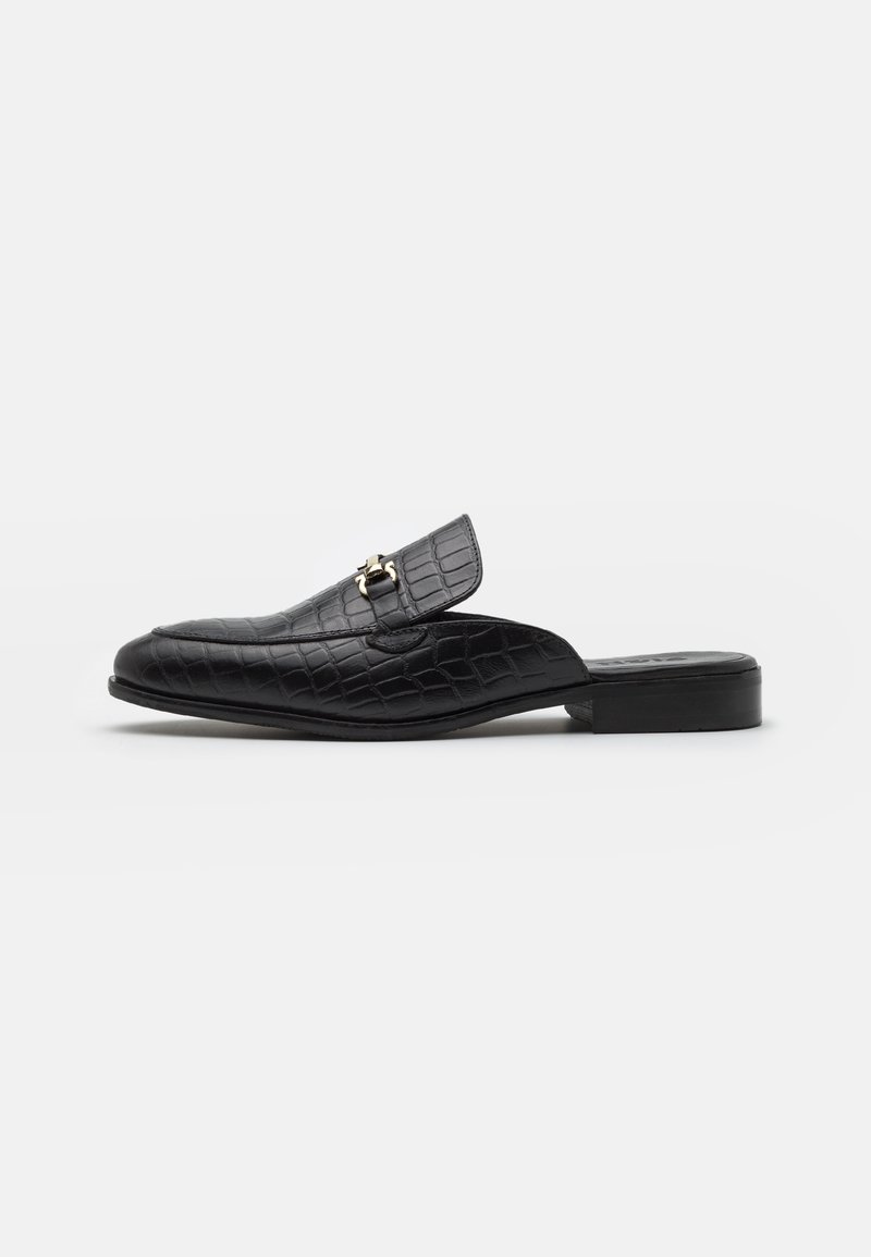 Sapatos slip-on de couro preto com padrão em relevo de crocodilo, com um salto plano e uma fivela metálica decorativa na tira. Textura suave e design elegante.