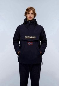 Jovem com cabelo encaracolado utiliza uma jaqueta preta com capuz, que possui um bolso frontal e um emblema da bandeira norueguesa.