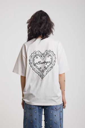 Pitkähiuksinen henkilö, jolla on valkoinen oversize-t-paita, jossa musta pitsisydän ja teksti "Darling", yhdistettynä sinisiin niittifarkkuihin, katsomassa poispäin.