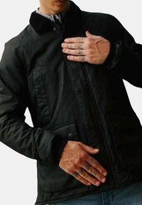 Giacca nera con colletto a revers, bottoni a pressione e tasche frontali doppie, abbinata a una camicia a quadretti sotto.