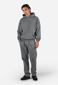 Young man wearing a gray hoodie and matching sweatpants with black sneakers, standing against a plain light background.
