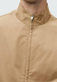 Beige zip-up jakke med høj krave, lavet af glat stof. Har en frontlynlås og minimalistisk design uden yderligere mønstre eller accenter.