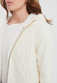 Giacca bianca a maglia con un design a trecce testurizzato, dotata di un cappuccio morbido e chiusura con zip, insieme a un top bianco a coste sottostante.