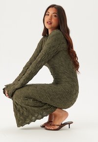 Olijfgroene jurk van volledige lengte met een gestructureerd ontwerp met verhoogde, herhalende patronen. Aansluitende silhouet met lange mouwen en uitlopende manchetten.