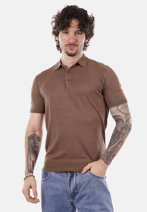 Uomo con capelli ricci che indossa una polo marrone a maniche corte e jeans blu, mostra tatuaggi su entrambi gli avambracci, in piedi con una mano in tasca.