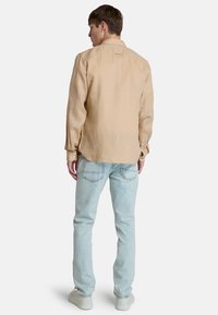 Camicia in lino beige chiaro a maniche lunghe con orlo leggermente arrotondato, abbinata a jeans in denim azzurro chiaro e sneakers bianche. Vestibilità casual.