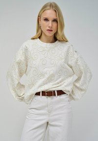 Pull blanc avec broderies florales dorées, coupe décontractée, manches larges. Associé à un jean clair et une ceinture marron.