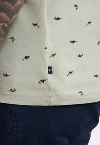 Světle zelená košile s malými, tmavě hnědými a zelenými vzory ryb; má černou nášivku na bočním švu; hladká bavlněná textura.