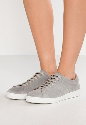 Coppia di piedi che indossano sneakers in suede grigio chiaro con suole e lacci bianchi, in piedi su un pavimento liscio e chiaro.