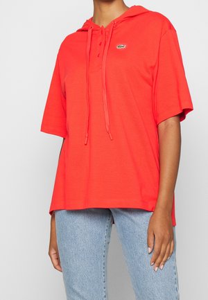 Osoba ubrana w jasnoczerwoną koszulkę z krótkim rękawem z kapturem i sznurkami oraz małym zielonym logo krokodyla, w połączeniu z jasnoniebieskimi dżinsami.