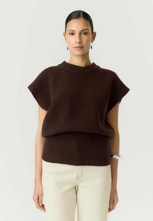 Femme aux cheveux foncés tirés en arrière, portant un haut en maille marron foncé à manches courtes et un pantalon beige clair, debout devant un fond clair uni.