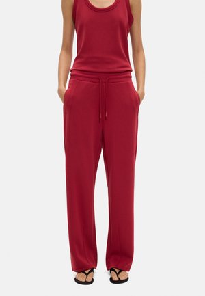 Szczupła osoba ubrana w czerwony bezrękawnik i luźne czerwone spodnie z troczkiem, ręce w kieszeniach, w czarnych klapkach.