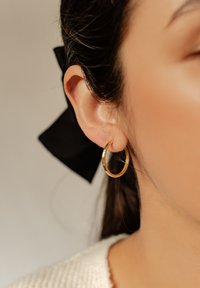 Goldener Creolenohrring, der an einem Ohrläppchen befestigt ist, mit glänzender Oberfläche. Eine schwarze Schleife im Haar ist sichtbar und hebt das Design des Ohrrings hervor.