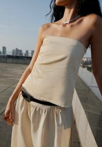 Haut beige sans bretelles avec un design froncé, tissu fluide et ourlet brut. Porté avec une ceinture noire munie d'une boucle rectangulaire en argent.