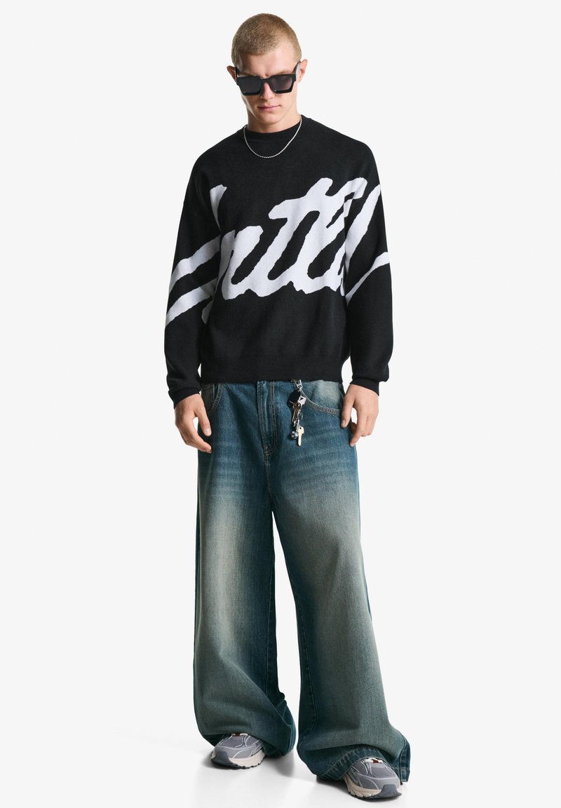 Black knit sweater with white cursive text, paired with oversized blue denim jeans, and gray sneakers. Accessories include a chain and keys.