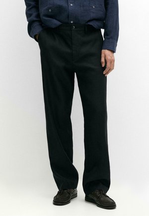 Homme portant un pantalon noir ample, une chemise bleu foncé boutonnée et des chaussures en cuir noires, debout avec une main dans la poche.
