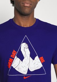 Sininen puuvillapaita, jossa on graafinen kuvasto koukistetuista käsivarsista kolmiomalli, sekä oranssia tekstiä "ARM DAY" ja "ME", lisäksi Nike-logo.