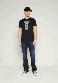 Μαύρο γραφικό T-shirt με κοντά μανίκια, που διαθέτει λευκό και μπλε σχέδιο, συνδυασμένο με μπλε τζιν παντελόνι και μαύρα αθλητικά παπούτσια.