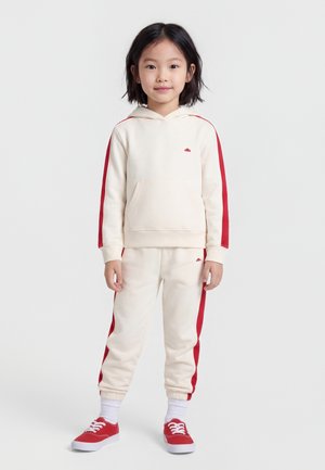 Νεαρή κοπέλα στέκεται φορώντας εκρού φούτερ με κουκούλα και παντελόνι με κόκκινες ρίγες, συνδυασμένα με λευκές κάλτσες και κόκκινα αθλητικά παπούτσια, μπροστά σε απλό φόντο.