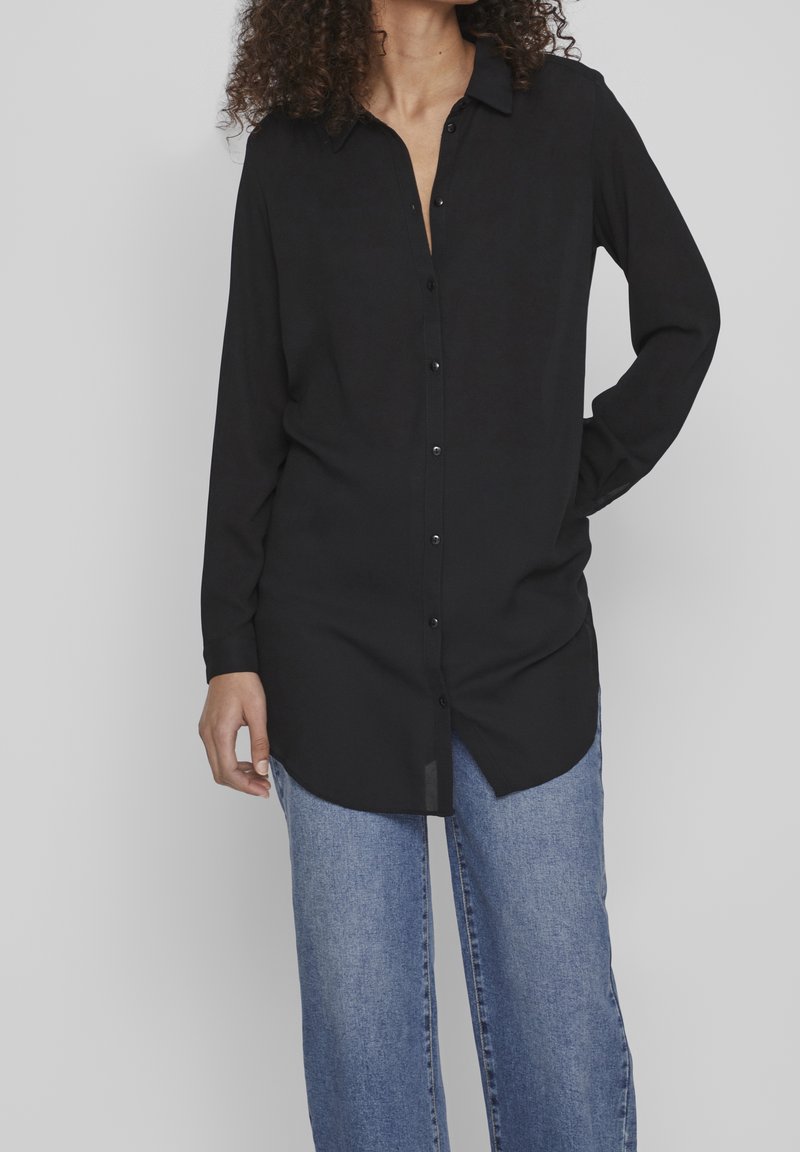 Personne portant une longue chemise noire boutonnée avec col et un jean droit bleu clair sur un fond uni.
