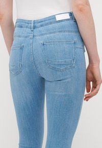 Jeans skinny de mezclilla azul claro con dos bolsillos traseros y una cinturilla recta, con una etiqueta en la parte posterior. Textura suave.