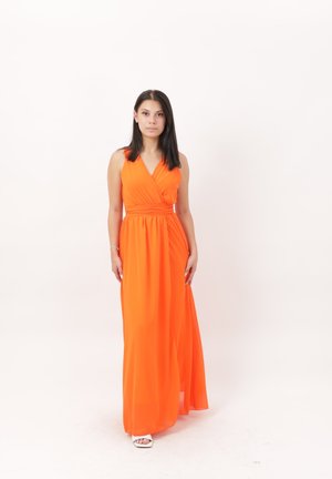 Femme aux cheveux raides et foncés, portant une robe longue orange vif sans manches, debout devant un fond blanc uni.