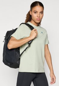 Crni ruksak od teksturiranog materijala, s podesivim trakama i srebrnim detaljima, uparen s svijetlozelenom majicom s malim plavim logotipom.