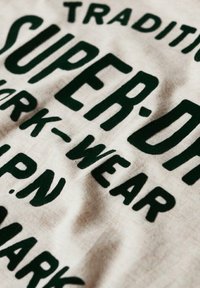 Cotton fabric with green bold text reading "SUPERDRY TRADITIONAL WORK-WEAR" on a light beige background, featuring a soft texture.