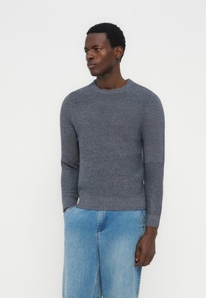 Hombre que lleva un suéter gris de punto acanalado con mangas largas y jeans azul claro, de pie con las manos en los bolsillos, mirando hacia un lado.