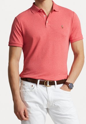 Homme portant un polo corail avec un petit logo brodé, un pantalon blanc, une ceinture en cuir marron et une montre-bracelet avec un cadran bleu et un bracelet marron.