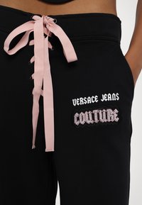 Zwarte sweatpants met een roze lintstrik, geborduurde "Versace Jeans" en "Couture" logo's, die de contrasterende kleuren en texturen benadrukken.