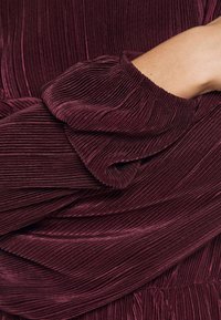 Burgunderfarbener, plissierter Stoff mit einem sanften Glanz, der eine geraffte, gerippte Textur und elastische Bündchen aufweist. Die Falten erzeugen Dimension und visuelles Interesse.