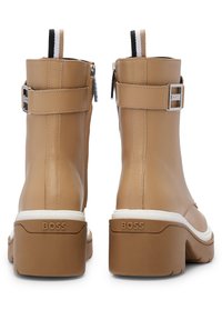 Botas de tobillo de cuero color marrón claro con un acabado brillante, que presentan un zipper lateral, correas ajustables y una suela blanca gruesa con marca.