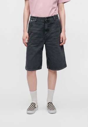 Person iført sorte knælange denimshorts, lyserød t-shirt, hvide strømper og sort-hvide ternede slip-on sko, stående mod hvid baggrund.