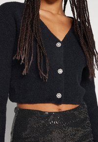 Musta lyhyt neuletakki teksturoidusta neuloskankaasta, V-kaula-aukolla ja koristenuppineuloilla, joissa on kukkakuosi.