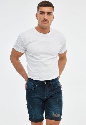 Hombre con cabello corto y oscuro y bigote, vestido con una camiseta blanca y pantalones cortos de mezclilla azul oscuro remangados, sobre un fondo blanco liso.