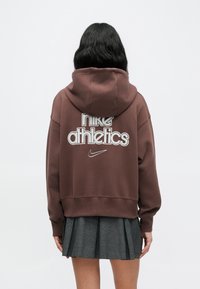 Καφέ φούτερ με κουκούλα και γράφικ "nike athletics" στην πλάτη. Ριμπ μανσέτες και κάτω μέρος, κατασκευασμένο από μαλακό, υφασμάτινο ύφασμα.
