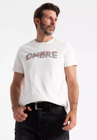 Camiseta de algodón blanca con el texto impreso "OMBRE" en negro y rojo. Mangas cortas, escote redondo y corte relajado. Se lleva con jeans negros.