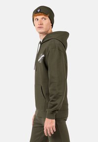 Felpa verde oliva con tasca frontale e stampa bianca del logo. Presenta un cappuccio foderato e polsini a coste. Indossata con un berretto coordinato.