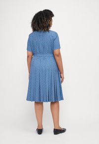 Mėlyna poliesterinė suknelė su baltais ir raudonais gėlių raštais, trumpomis rankovėmis, klostuotu sijonu ir juosmeniu, kuris yra sutrauktas. Dėvima su juodomis butais.