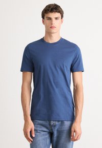 Joven con pelo corto y rizado, vistiendo una camiseta azul lisa de manga corta y jeans de color azul claro, de pie contra un fondo blanco.