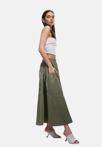 Gonna lunga verde oliva con tasche laterali arricciate e cintura elastica, abbinata a un top corto bianco e mules aperti grigi chiari.