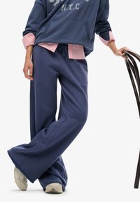 Persona vistiendo pantalones de chándal de pierna ancha azul marino, zapatillas grises claras, una sudadera azul marino sobre una camiseta rosa, descansando la mano sobre una silla de madera oscura.